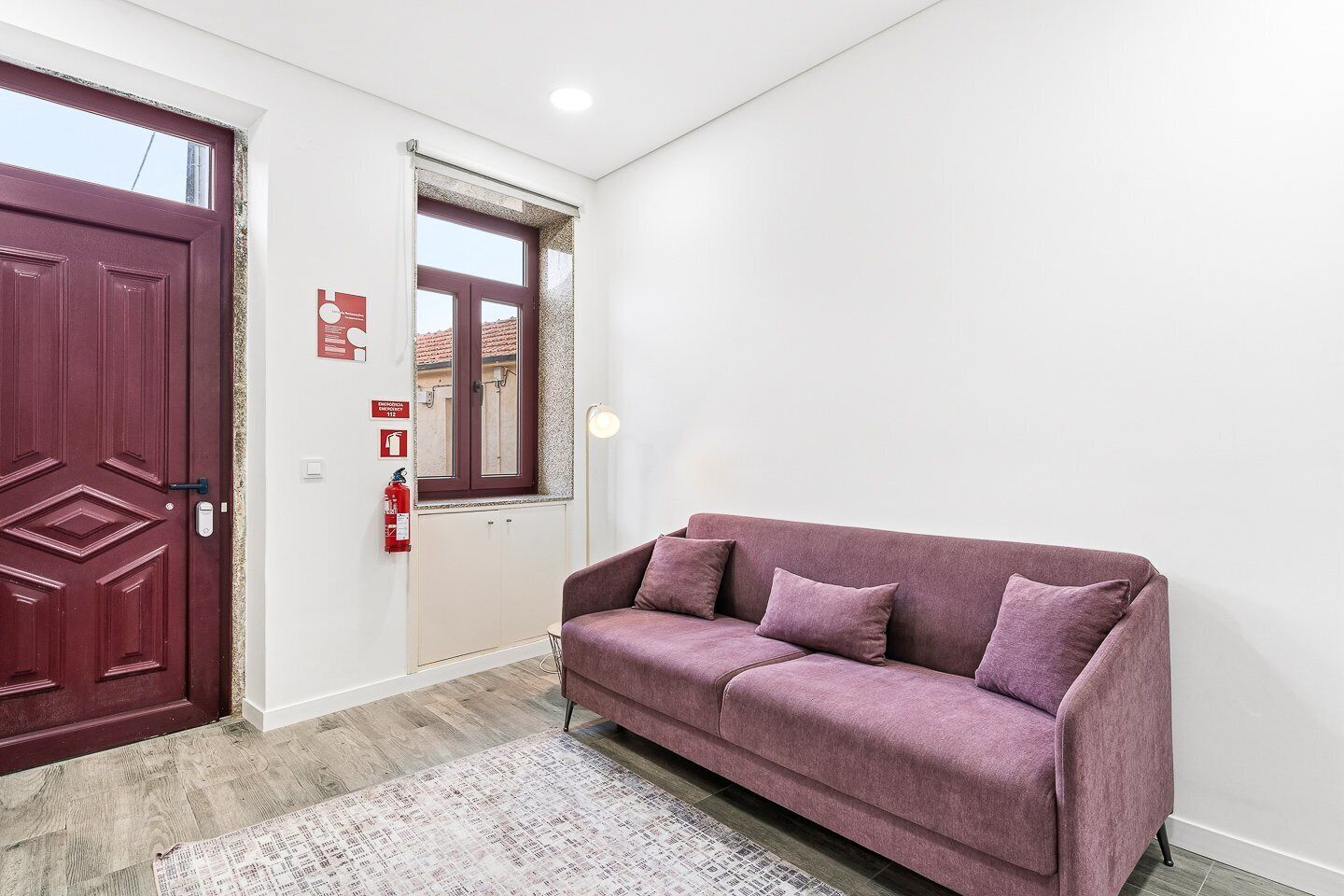 Ein Zimmer mit roter Tür, einem Fenster mit Ausblick, einem violetten Sofa und einem Feuerlöscher an der Wand.