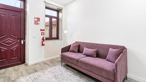 Ein Zimmer mit roter Tür, einem Fenster mit Ausblick, einem violetten Sofa und einem Feuerlöscher an der Wand.
