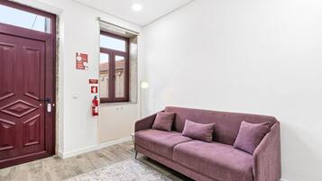 Ein Zimmer mit roter Tür, einem Fenster mit Ausblick, einem violetten Sofa und einem Feuerlöscher an der Wand.