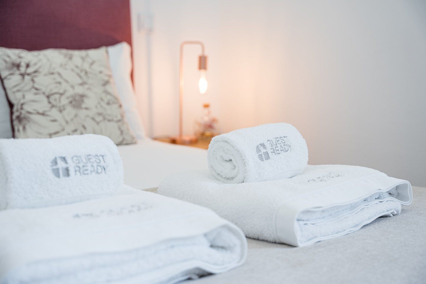 En pænt redt seng med et 'Guest Ready' håndklæde og en lampe i baggrunden.