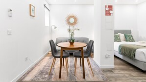 Et moderne værelse med spiseplads, der inkluderer et rundt bord, grå polstrede stole og et dekorativt spejl. En seng med en pude med bladmotiv er synlig i baggrunden.
