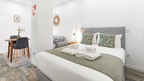 Et moderne hotelværelse med en pænt redt seng, en grå polstret stol, et lille bord med en vase med blomster og et dekorativt spejl på væggen.