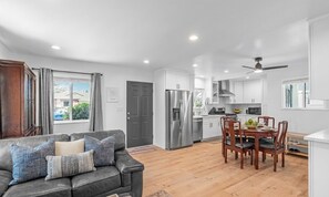 A modern kitchen with white cabinets, a stainless steel refrigerator, and a dining area with wooden chairs and a table. - New 2BR/2BA Furnished Home w/Yard LAX/SoFi/Intuit (Inglewood)