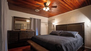 A bedroom with a wooden ceiling, a bed with a headboard, a bench, a dresser, a mirror, and a ceiling fan.