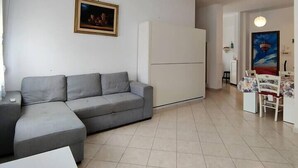 A modern living room with a grey sectional sofa, a white coffee table, and a dining area with a floral tablecloth. - Cozy Flat Near Beach | WiFi & Comfort, Torvaianica (Torvaianica)