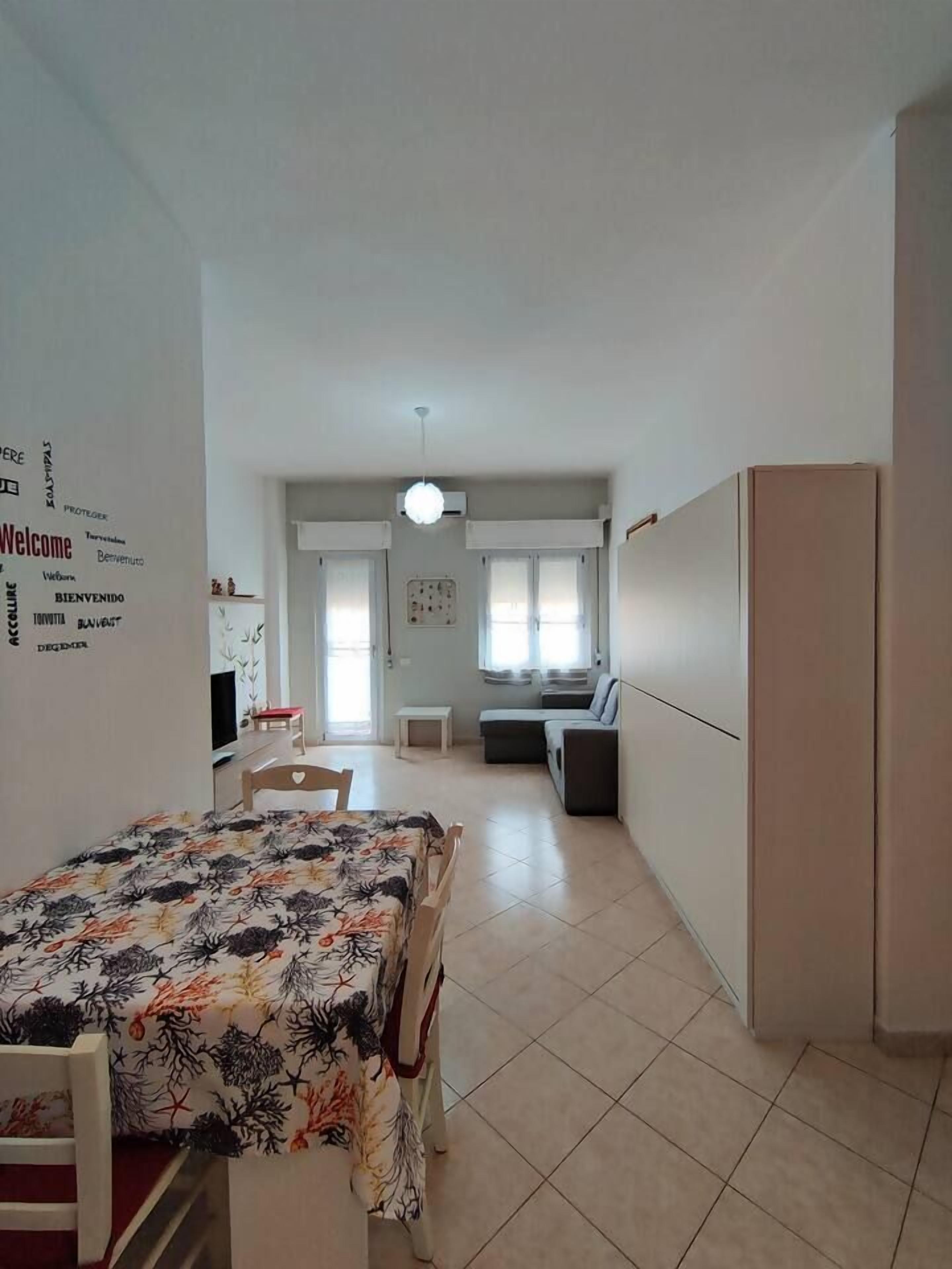 A room with a bed, a desk, a chair, a small table, and a sofa. There is a window with curtains, a wall-mounted air conditioning unit, and a door leading to another room.