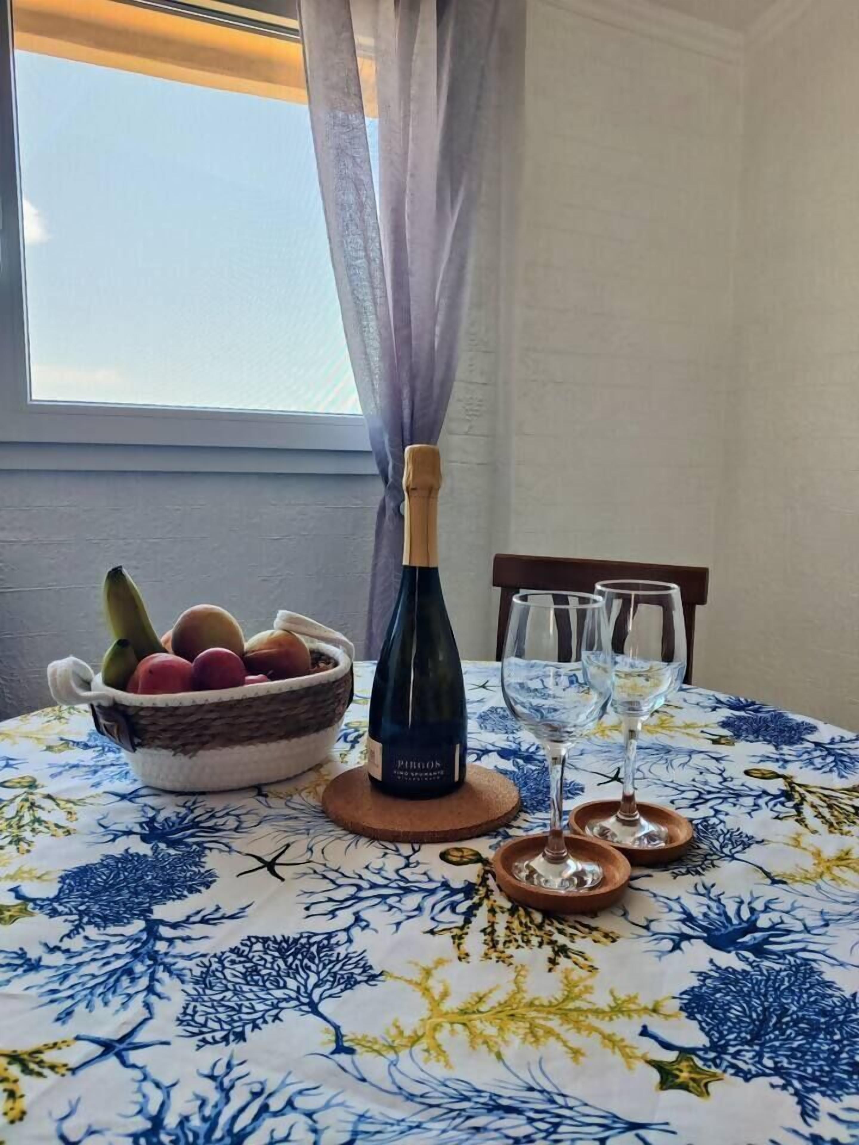 Ein Esstisch mit einer blumigen Tischdecke, einer Weinflasche, einer Obstschale und zwei Weingläsern.