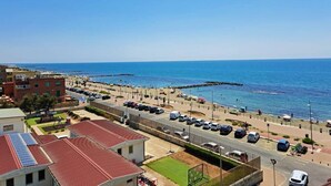 Vista costera con una carretera que lleva a una playa, coches estacionados y edificios con tejados rojos.