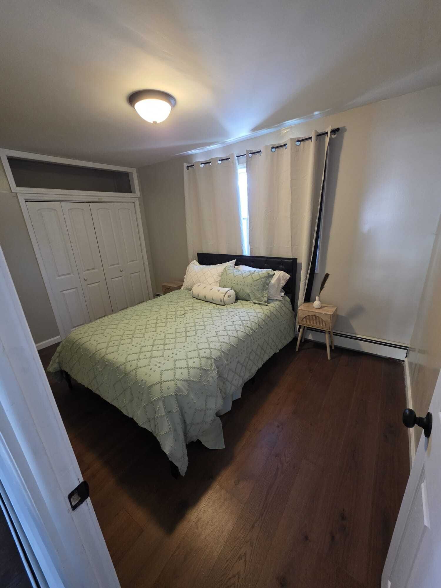 Une chambre à coucher avec un lit, une table de chevet, une fenêtre avec des rideaux, un luminaire au plafond et une porte.
