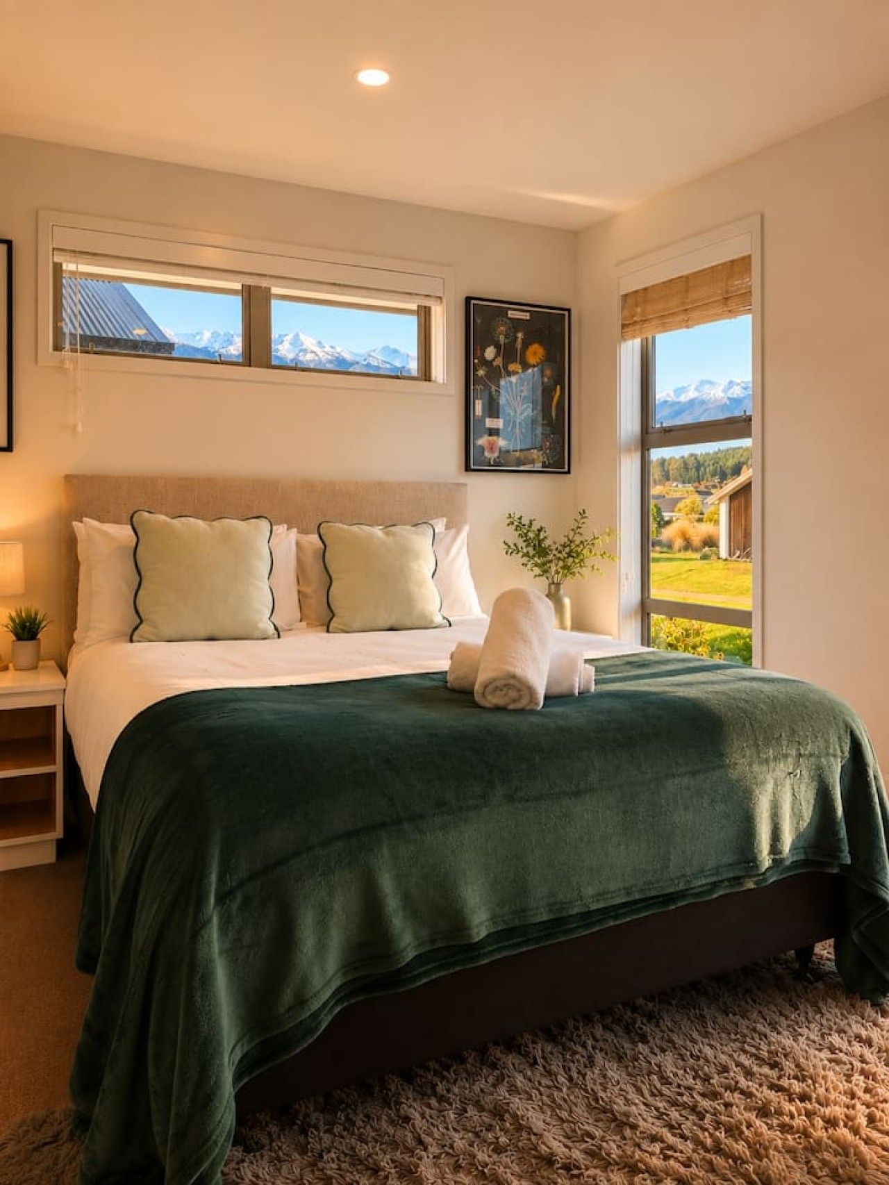Une chambre à coucher avec un lit, des oreillers, une couverture verte, une fenêtre donnant sur les montagnes et un tableau encadré accroché au mur.