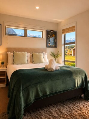 A bedroom with a bed, pillows, a green blanket, a window with a view of mountains, and a framed picture on the wall. - Shotover Sanctuary - 2 Bedroom home! (Queenstown)