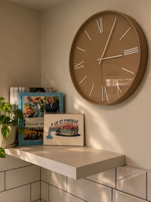 Une horloge murale avec des chiffres romains, un livre encadré intitulé « Une gorgée de paradis », et une plante en pot posée sur une étagère.