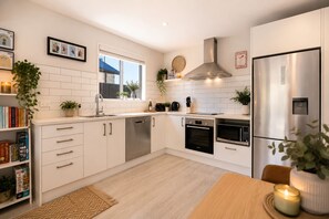 A modern kitchen with white cabinetry, stainless steel appliances, a window, and a dining area with a table and chairs. - Shotover Serenity (Queenstown)