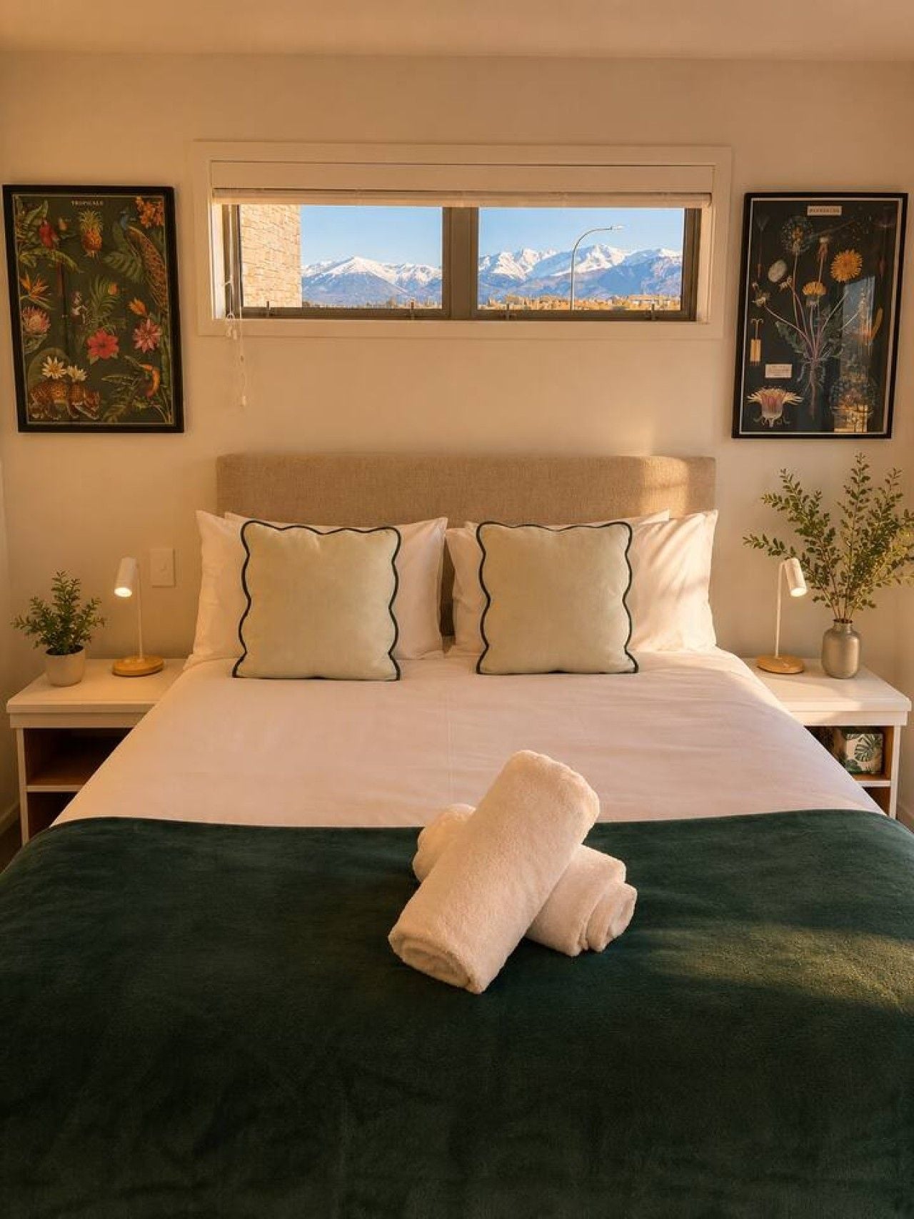 Una cama con vistas a las montañas a través de una ventana, dos mesitas de noche con lámparas e impresiones enmarcadas de plantas en las paredes.