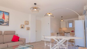 A modern living room with a glass coffee table, a beige sofa with a red pillow, and a dining area with a white table and clear chairs. There is a kitchen in the background with white cabinets and a refrigerator. - 4-STAR APARTMENT – 2 BEDROOMS – COGOLIN – SWIMMING POOL – CLOSE TO THE TOWN CENTRE (COGOLIN)
