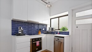 A modern kitchen with white cabinets, a blue-tiled backsplash, a built-in oven, a dishwasher, and a window with a plant outside. - Relaxing Stay Near Fairlight's Scenic Spots (Sydney)