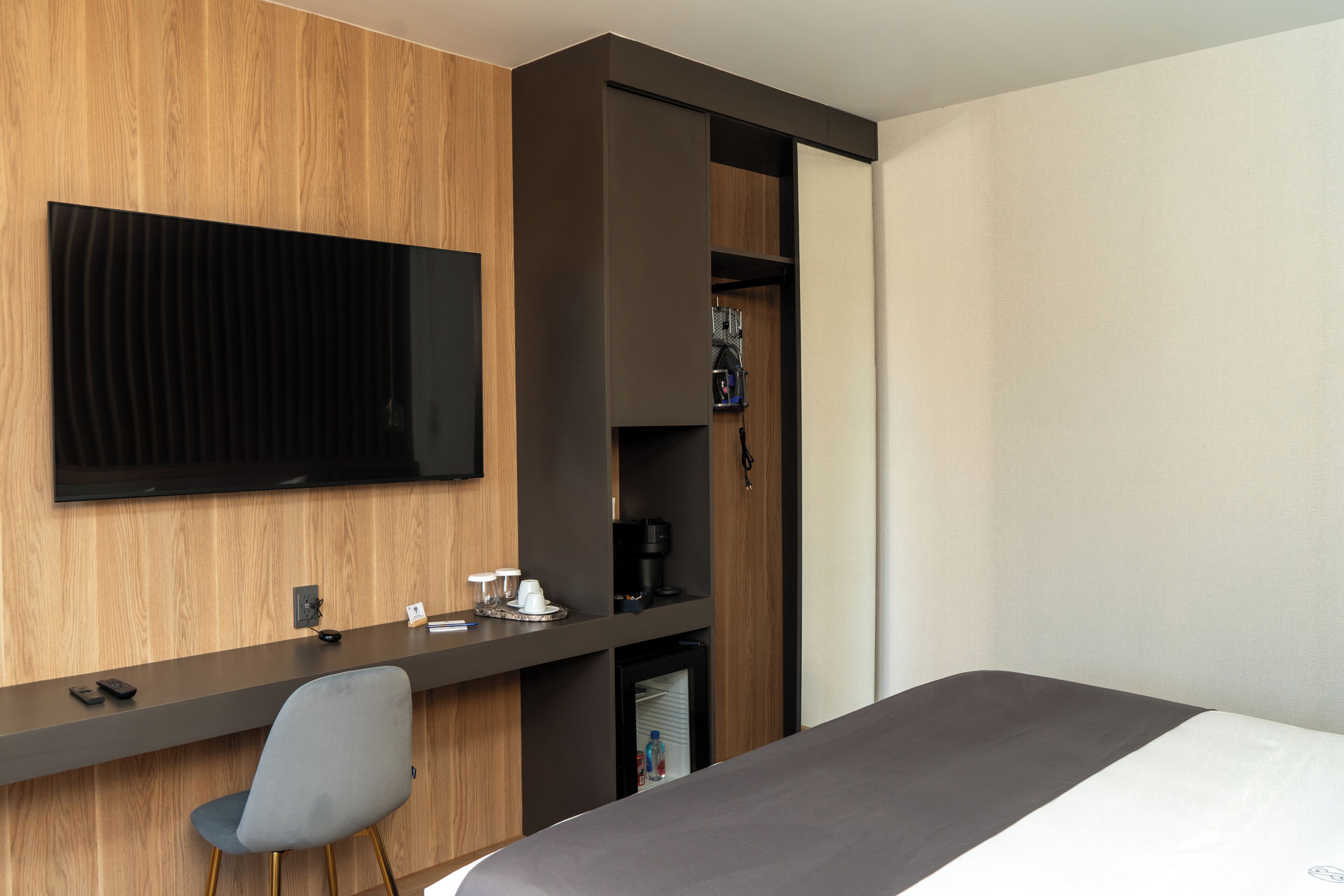 A hotel room with a bed, a desk with a chair, a television mounted on the wall, and a minibar.