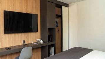 A hotel room with a bed, a desk with a chair, a television mounted on the wall, and a minibar.