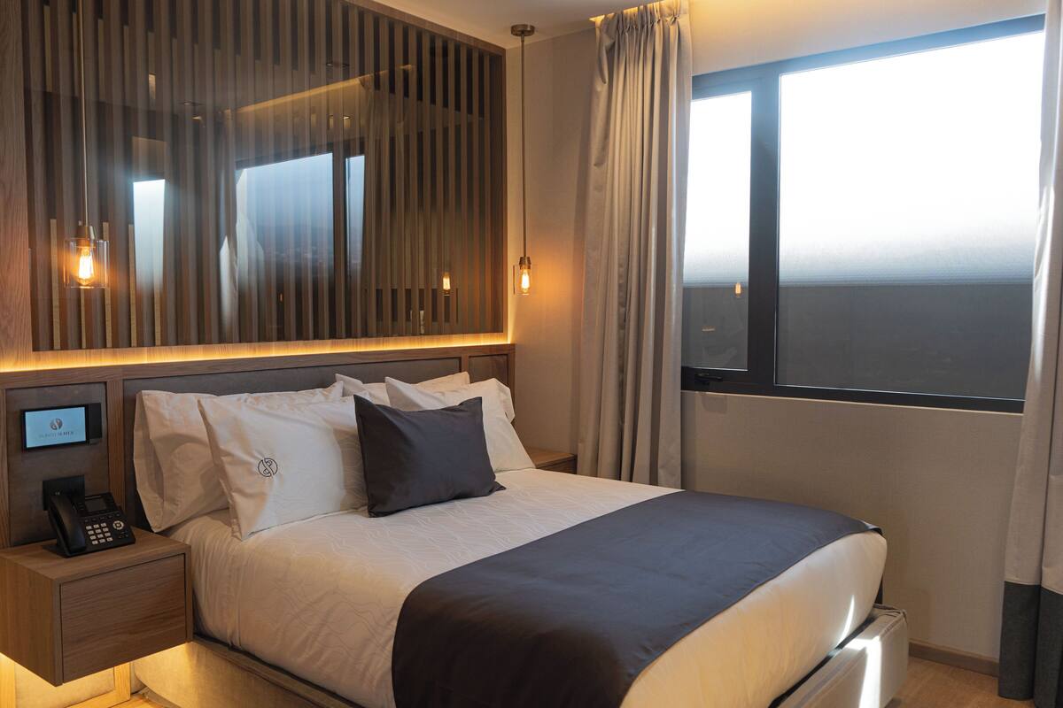 Habitación de hotel con cama, mesita de noche con teléfono, espejo y ventana con cortinas.