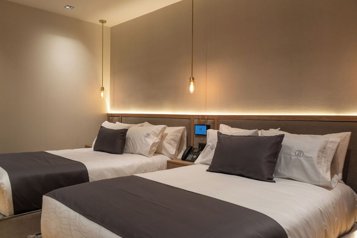 Quarto de hotel com duas camas, uma mesa de cabeceira com telefone e televisão presa à parede.