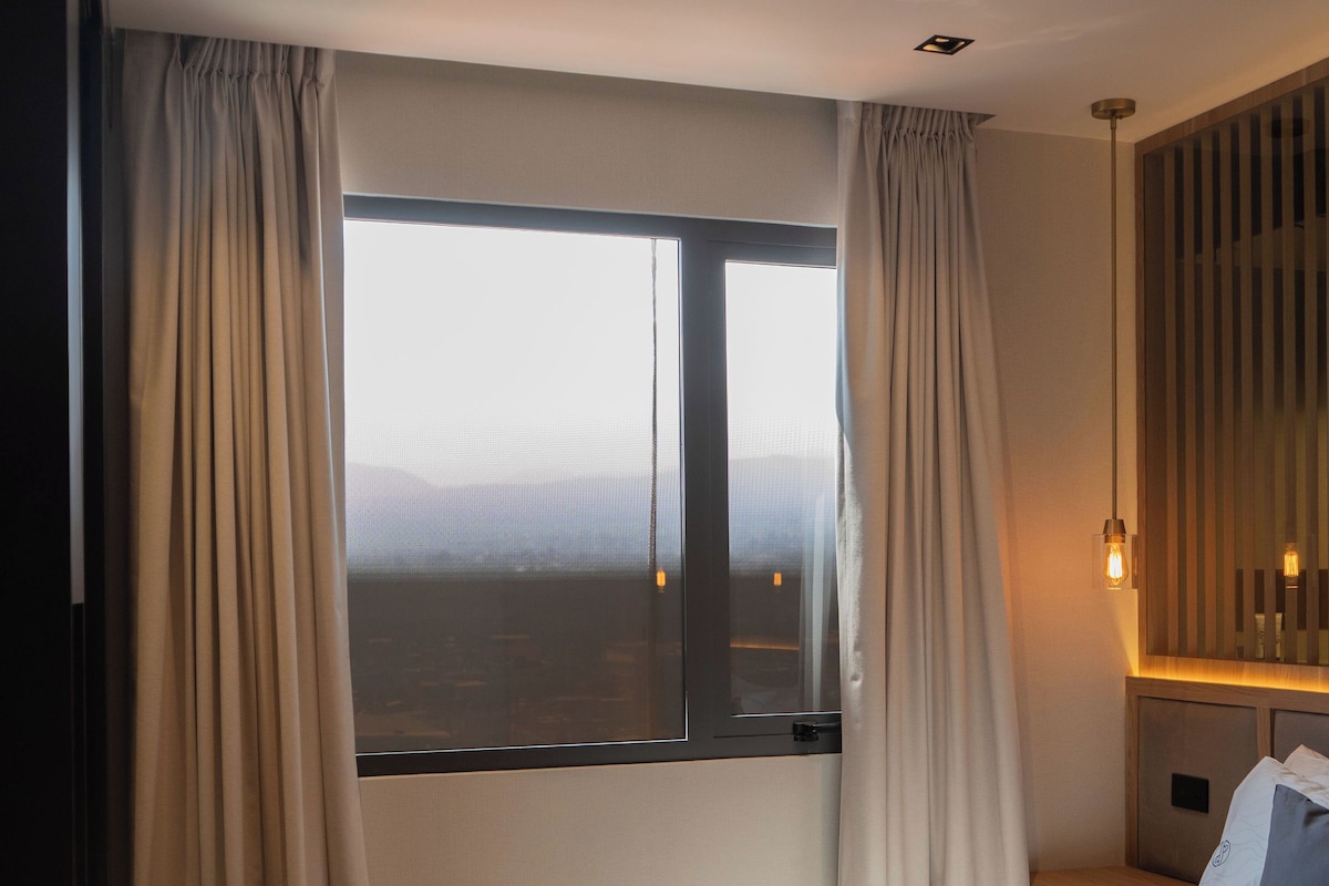 Quarto de hotel moderno com uma cama grande, vista pela janela e mesa de cabeceira com abajur.