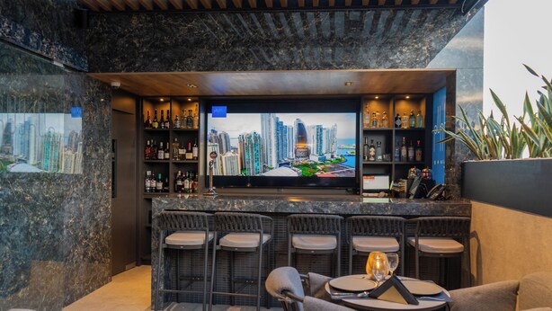 Un'area bar con TV che mostra un panorama urbano, sgabelli da bar e un'area salotto con tavoli e sedie.