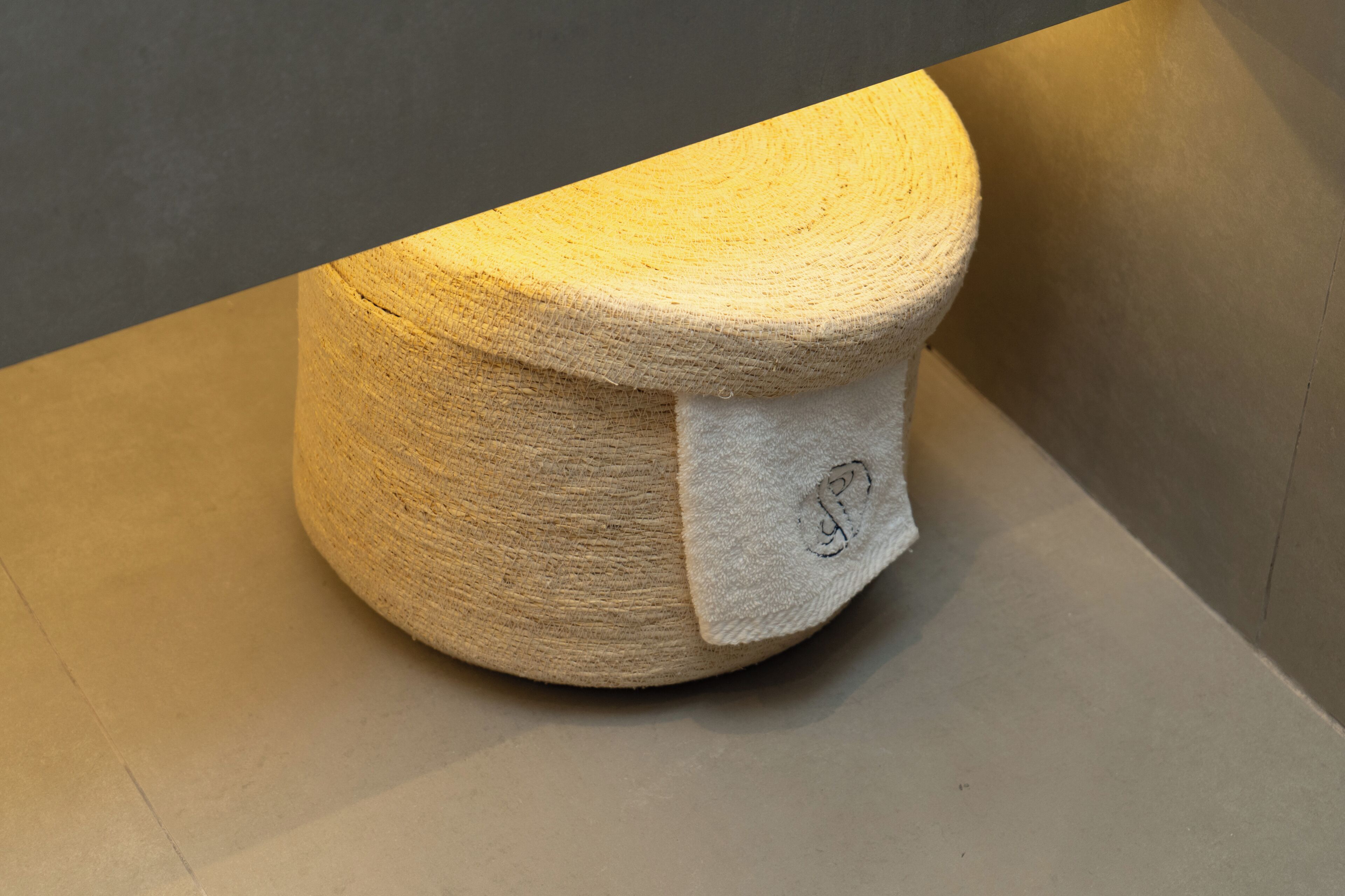Ein runder, strukturierter Hocker mit weißem Sitzkissen und kleinem eingesticktem Logo.