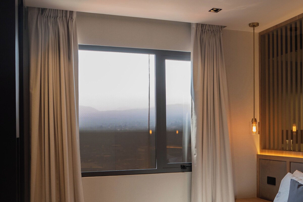 Una habitación de hotel moderna con una cama grande, vistas a través de una ventana y una mesita de noche con lámpara.