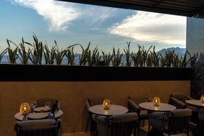 Een restaurant met ronde tafels en stoelen, uitzicht op bergen en een rij planten aan de bovenkant.
