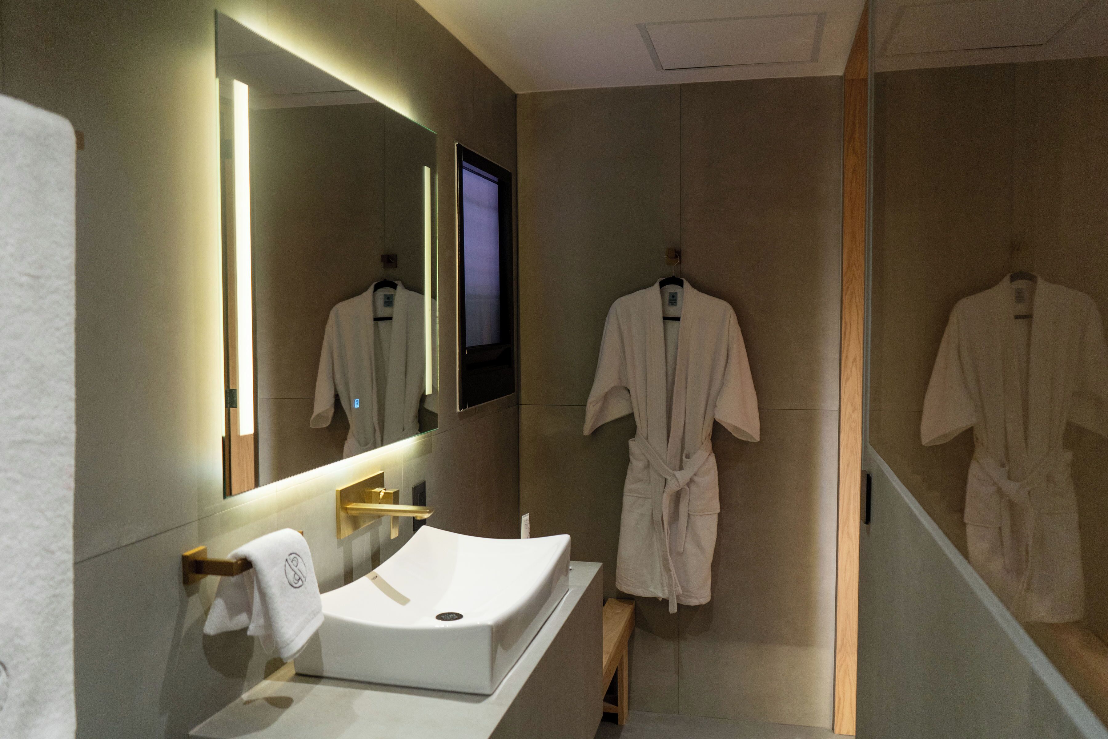 Une salle de bain moderne avec un lavabo blanc, un grand miroir et deux peignoirs blancs suspendus à des étagères en bois.