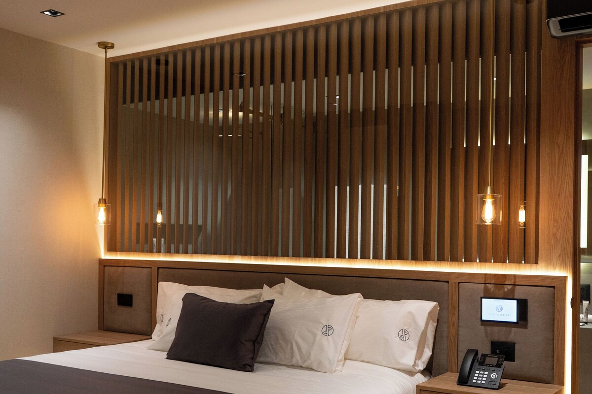 Ein modernes Hotelzimmer mit einem großen Bett, einem Holz-Kopfteil, Nachttischen mit Lampen und einem integrierten Fernseher.