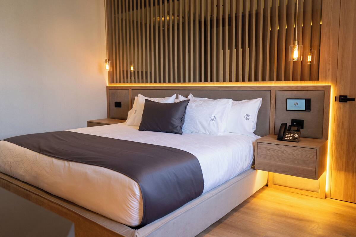 Una habitación de hotel moderna con una cama grande, cabecera de madera, mesitas de noche y un teléfono sobre una de ellas.