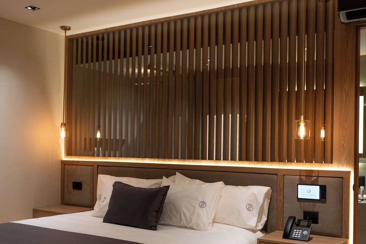 Habitación de hotel moderna con una cama grande, cabecera de madera, mesitas de noche con lámparas y televisión integrada.