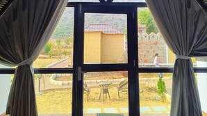 A room with a large window framed by curtains, offering a view of a courtyard with outdoor seating and a building in the background.