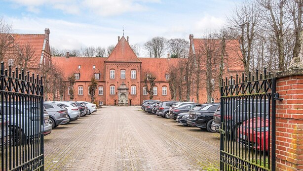 A paved driveway leading to a brick building with a red roof, surrounded by a metal fence and parked cars. - 4 Star Holiday Home in Skanderborg (Skanderborg)