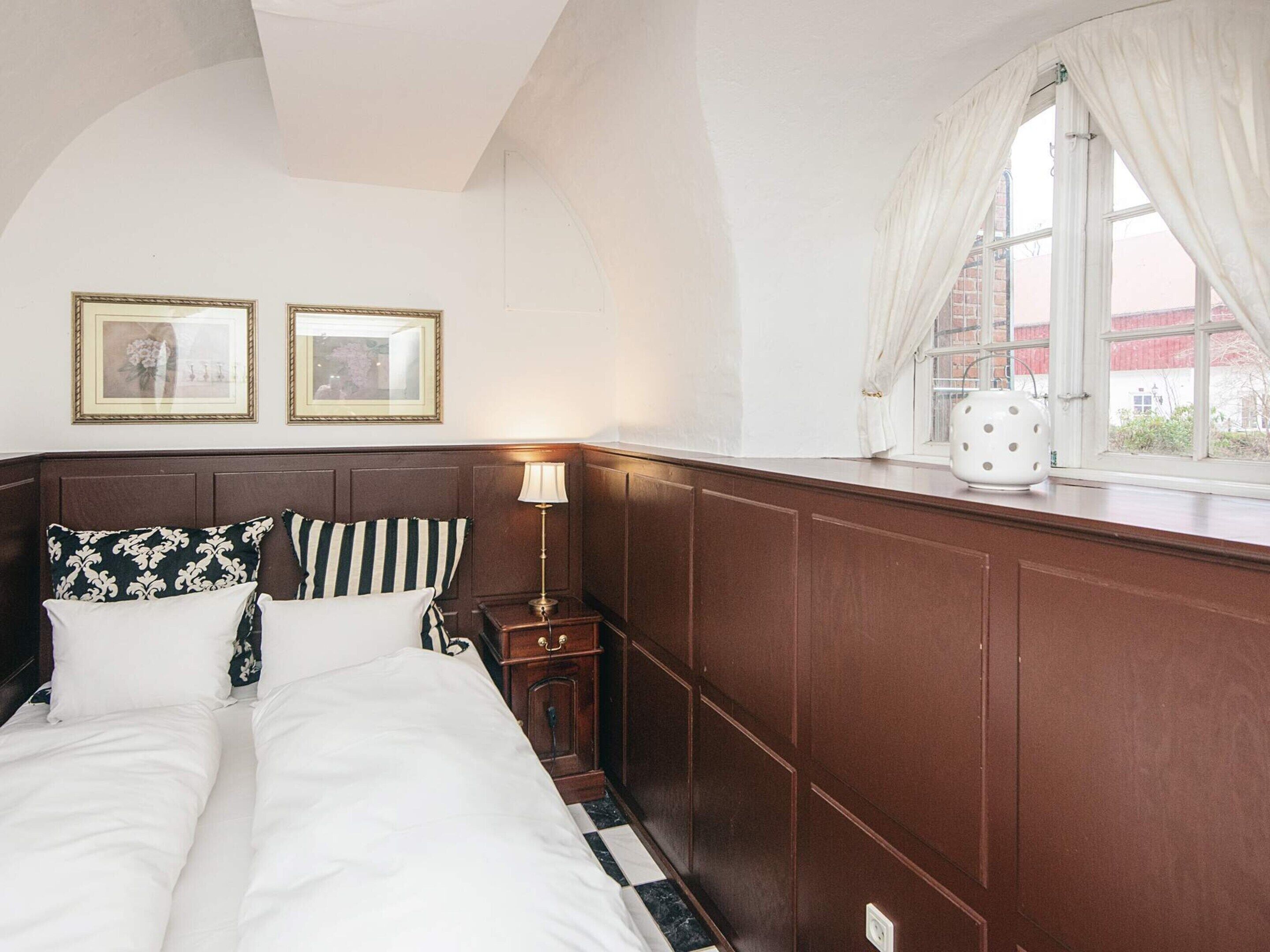 A bedroom with a large bed, two framed pictures on the wall, a bedside table with a lamp, and a window with curtains.