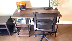 Ett skrivbord i trä med en svart stol med nätmaterial, en inramad bild av en solnedgång och en liten krukväxt.