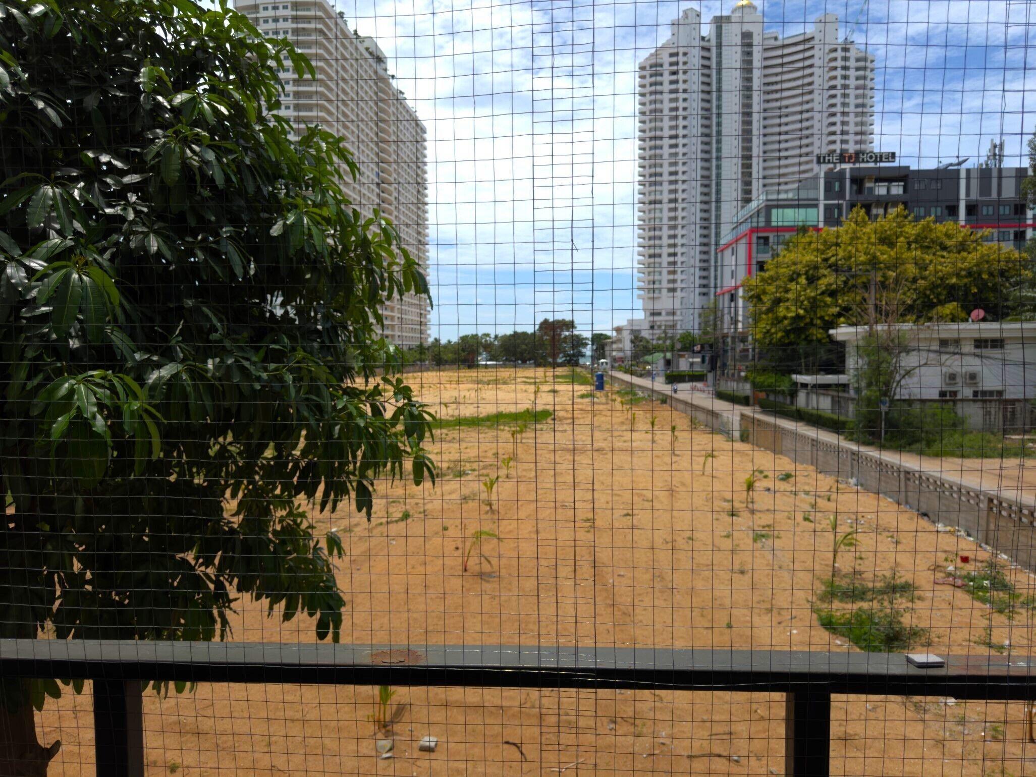 Una zona arenosa con una cerca, edificios al fondo y un árbol a la izquierda.