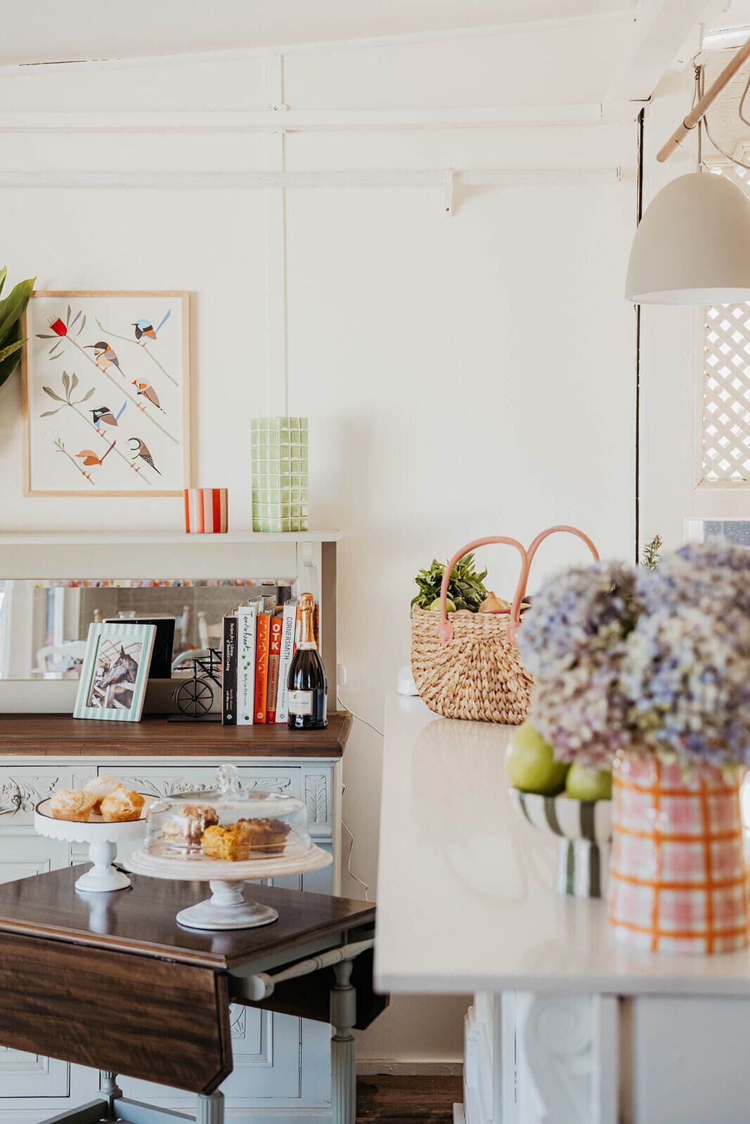 A kitchen with a wooden countertop, a white cabinet, and a hanging light fixture. There is a framed artwork on the wall and a basket of flowers on the counter.