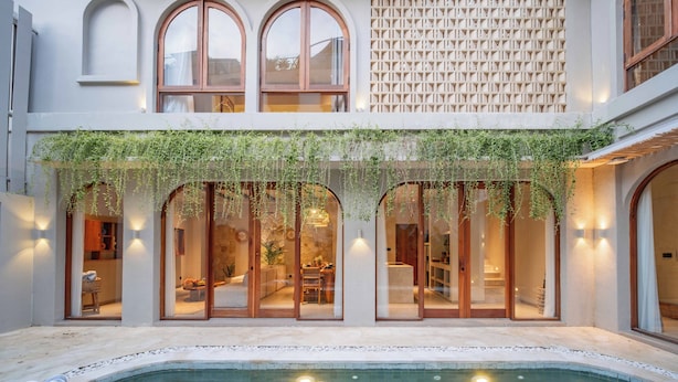 Área à beira da piscina com espreguiçadeira, banheira de hidromassagem e vista do interior através de grandes janelas em arco.