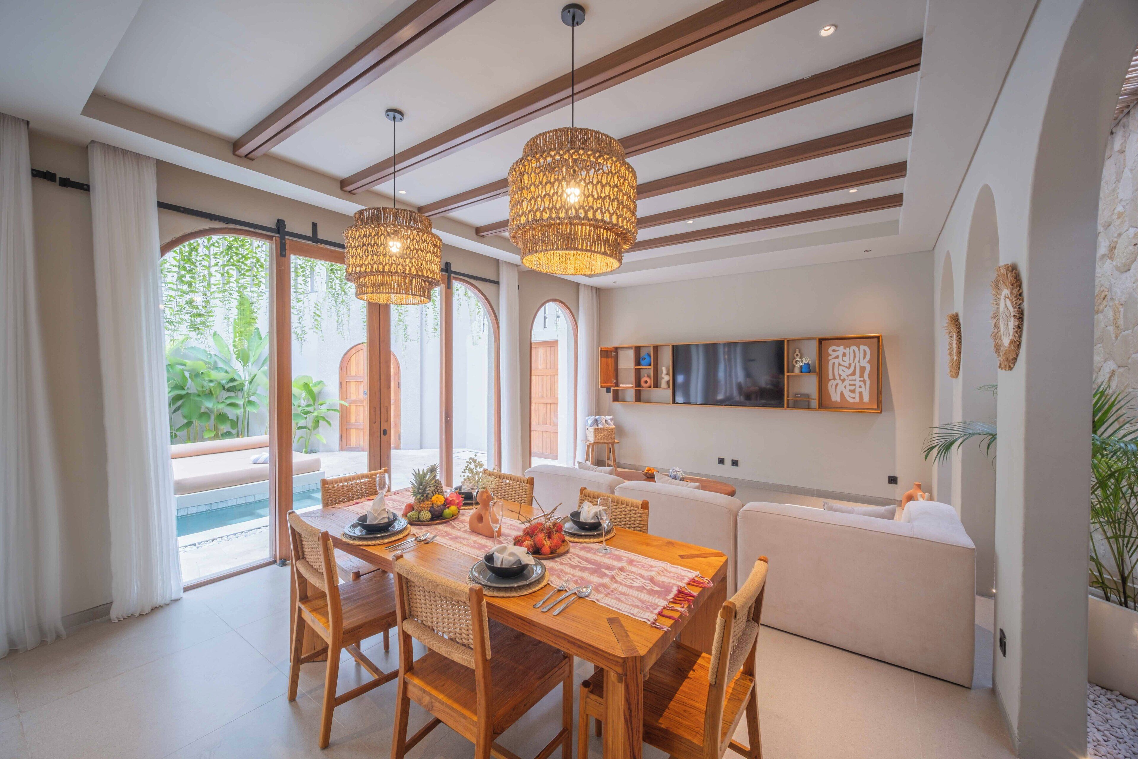 A spacious dining area with a wooden table, wicker chairs, and hanging wicker lamps. There is a large window with curtains and a view of greenery outside.