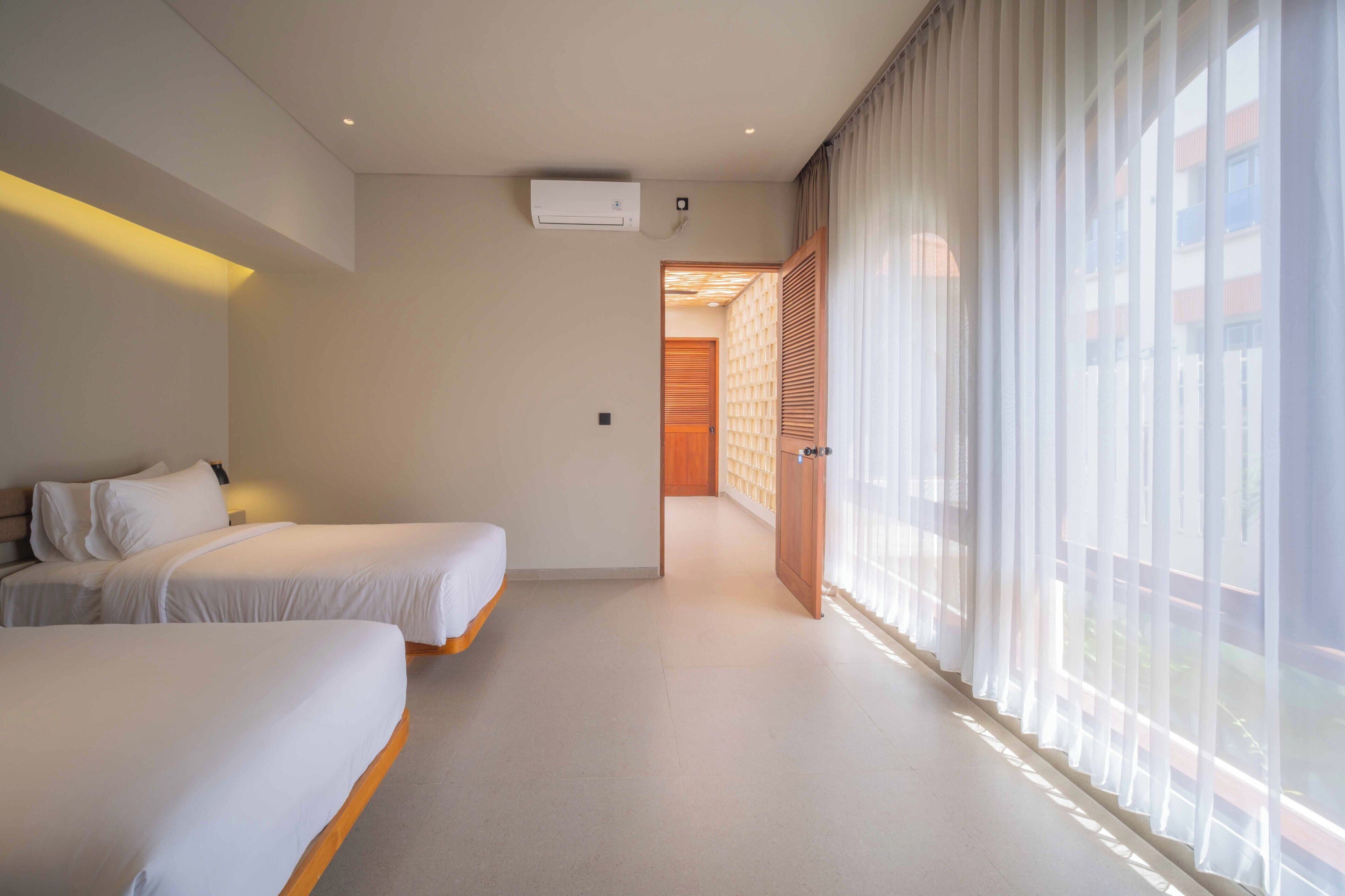 Una habitación de hotel con dos camas, un ventanal con cortinas translúcidas y una puerta de madera que da a otra habitación.