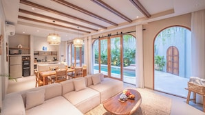 Uma sala de estar ampla com um sofá grande, uma mesa de centro de madeira e uma mesa de jantar com cadeiras. Cozinha visível ao fundo, e grandes janelas oferecem vista para o exterior.