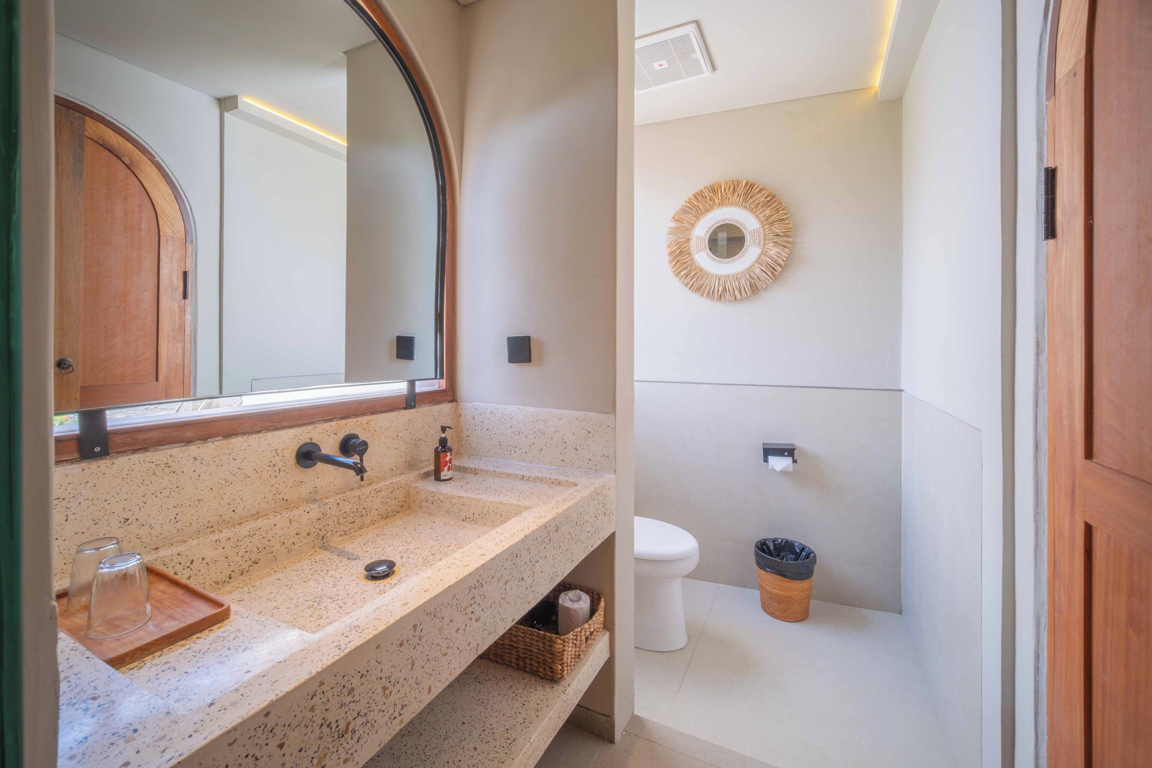A bathroom with a granite sink, a wooden door, a mirror, a toilet, a trash bin, and a wall-mounted towel rack.
