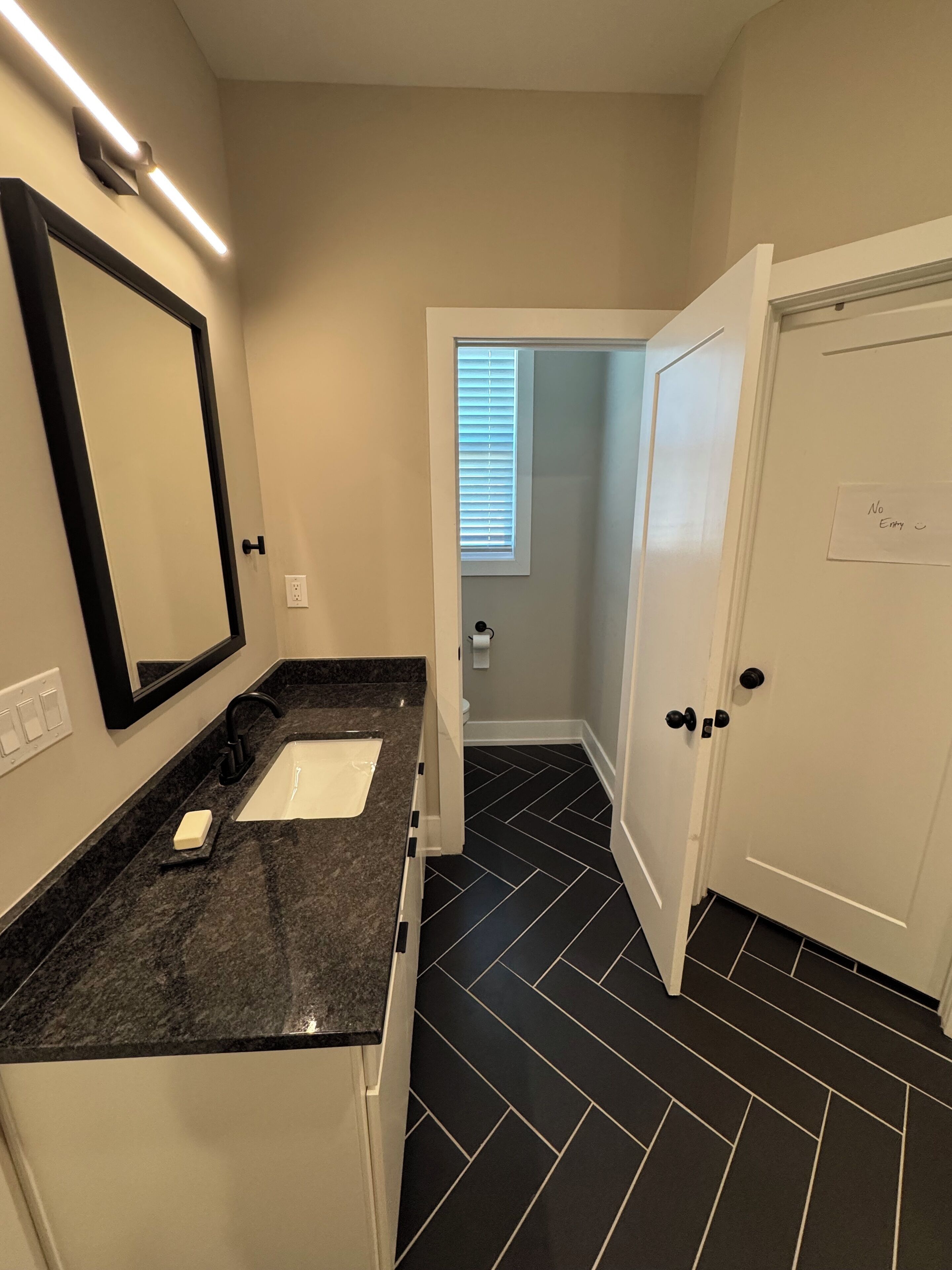 Une salle de bain avec un lavabo blanc, un plan de toilette noir et un grand miroir. Il y a une porte sur laquelle est apposée une pancarte indiquant « Entrée interdite ». Le sol est recouvert de carreaux noirs.