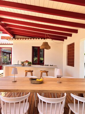 A dining area with a wooden table, white chairs, a hanging light fixture, and a window with a view of greenery. - Charm and tranquility under the maritime pines, stone's throw from the beach (Marina di Campo)