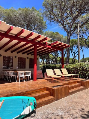 A covered outdoor seating area with wooden tables and chairs, a lounge chair, and a paddleboard. - Charm and tranquility under the maritime pines, stone's throw from the beach (Marina di Campo)
