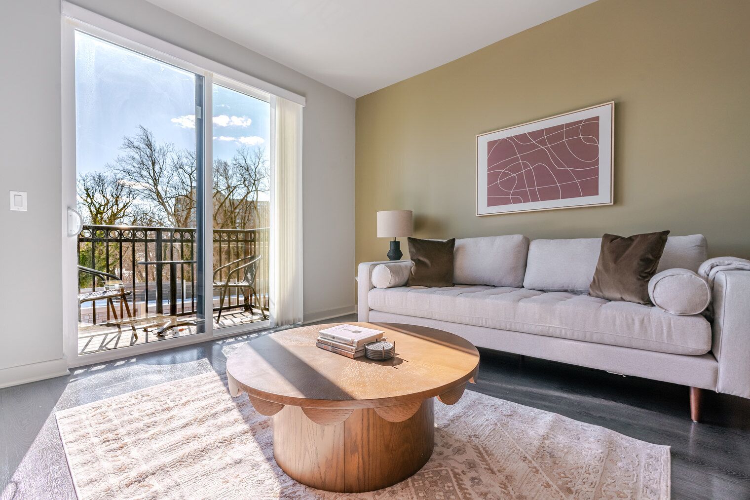 Een woonkamer met een grote schuifpui die uitkomt op een balkon, een ronde houten salontafel, een beige bank en een lijstwerk abstract kunstwerk aan de muur.