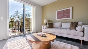 Een woonkamer met een grote schuifpui die uitkomt op een balkon, een ronde houten salontafel, een beige bank en een lijstwerk abstract kunstwerk aan de muur.