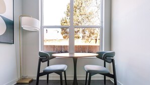 A corner room with a round table and two chairs, a floor lamp, and a window with a view of a wooden fence and trees. - Blueground | Chevy Chase, rooftop, nr Rock Creek Park (Chevy Chase)